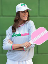 A model stands with her arms crossed holding a pickleball paddle. Her sweatshirt says "Pickleballer" in green and pink letters, and she has a matching white ball cap that is embroidered to say "Baller" with icons of pickleball paddles below