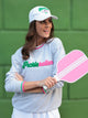 A model stands with her arms crossed holding a pickleball paddle. Her sweatshirt says 