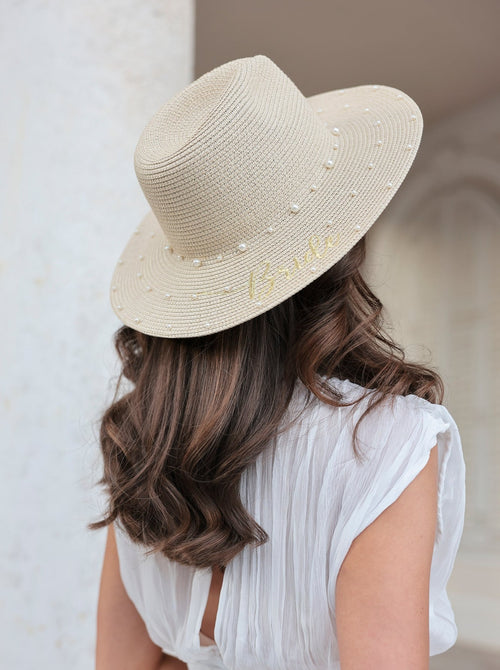 A model faces away from the camera to show a naturally colored woven hat with faux pearl embroidery and the word 